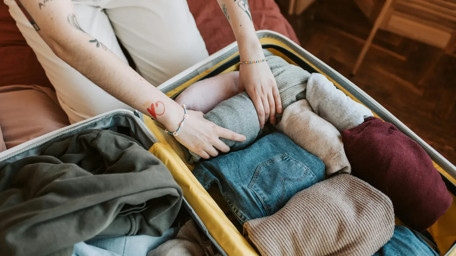 Hands rolling clothes into an open suitcase on a bed: blue jeans, gray, beige and burgundy knits, with an olive hoodie in the lid